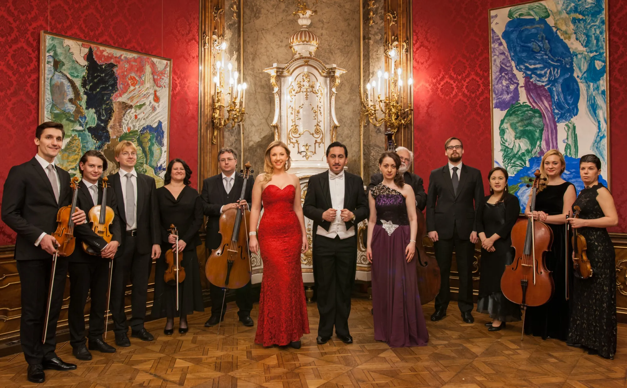 The Wiener Barock Orchester standing with their instruments before the red damask and gilt sconces of the Roter Salon, the soprano in red at the centre.